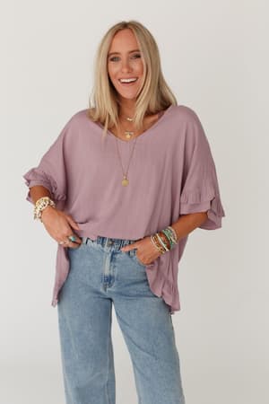 A woman with long blonde hair smiles while wearing The Nest Let Me Be Ruffled Top in Pale Plum, styled with light blue jeans, layered necklaces, and colorful bracelets, standing against a plain white background.