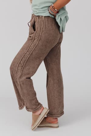 Wearing the So Comfy Wide Leg Pant Full Length - Mocha, a person with only their lower half visible pairs the loose, high-rise pants with a light green top, beaded bracelets, and beige espadrille sandals against a plain light background.