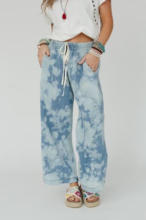 A person wearing The Nest So Comfy Wide Leg Denim Pant - Bleach Dye Denim, a white t-shirt, colorful bracelets, rings, and multicolor platform sandals stands against a plain white background.