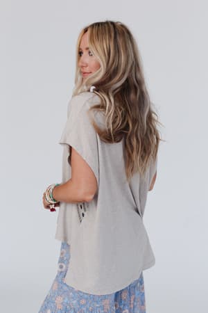 A woman with long, wavy blonde hair stands sideways, looking over her shoulder. She wears a loose Camden Lace V Neck Top in Camel, a blue floral skirt, and beaded bracelets against a plain light background.