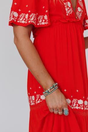 A person in a bright red dress with white floral embroidery is shown from shoulders to knees. Their hand rests by their side, adorned with silver rings and a layered bracelet from the San Antonio Bracelet Set - Turquoise.