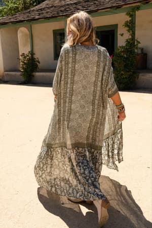 A woman with wavy blonde hair, seen from behind, wears the Paisley Tapestry Free Flow Duster Kimono in Olive and wedge sandals as she strolls outside near a rustic building with green-trimmed windows and flowering bushes.