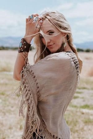 A woman with long blonde hair wears the Dreamy Macramé Kimono in Mocha, featuring tassel fringe and boho style, paired with statement jewelry as she stands outdoors in a grassy field under a bright sky, looking at the camera.