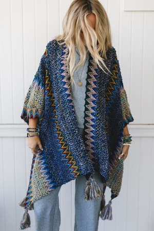 A person with long, wavy blonde hair wears the Desert Dreams Crochet Ruana - Cobalt, featuring colorful zigzag patterns and tassels over a light blue outfit, standing against a white paneled backdrop—ideal for any boho closet.