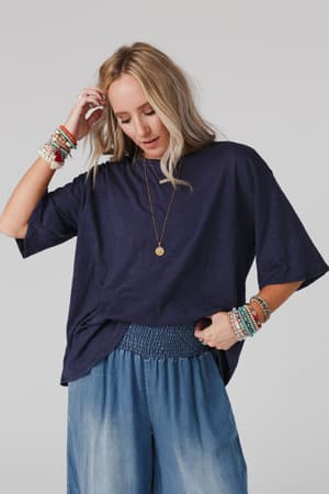 A woman with long blonde hair models The Nest Your Favorite Slub Tee Shirt in navy, styled with wide-leg denim pants, layered bracelets, and a long necklace, posing with one hand in her hair against a light background.