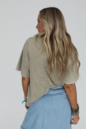 A woman with long, wavy blonde hair faces away, wearing the Fly Free Graphic Tee in mocha and a light blue denim skirt. She accessorizes with several bracelets on her right wrist against a plain light gray background.