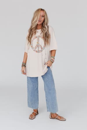 A woman with long, wavy blonde hair wears the Jude Oversized Peace Sign Graphic Tee in oatmeal, paired with light blue wide-leg jeans, sandals, and colorful bracelets, standing against a plain white background.