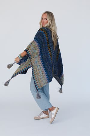 A woman with long blonde hair models the Desert Dreams Crochet Ruana in Cobalt, featuring a vibrant zigzag knit pattern, as she poses against a white background with boho-chic style.