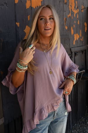 A woman with long blonde hair wears The Nest Let Me Be Ruffled Top in pale plum and blue jeans, accessorized with multiple bracelets, rings, and layered necklaces as she stands before a weathered dark wall with peeling orange paint.