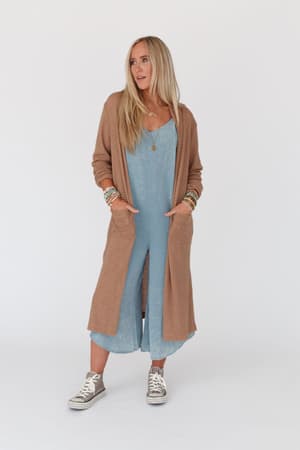 A woman with long blonde hair wears a light blue dress, gray high-top sneakers, layered bracelets, and The Nest Jayde Waffle Knit Cardigan in taupe, standing against a plain white background with her hands in her pockets.