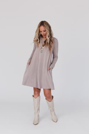 A woman in The Nest Jake Long Sleeve Crewneck Dress - Mocha Latte stands against a plain backdrop, wearing white knee-high boots, layered necklaces, and has long wavy blonde hair. She looks down with her hands in the dress pockets.