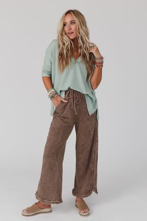 A woman with long wavy blonde hair wears a mint green loose-fit top, So Comfy Wide Leg Pant Full Length in Mocha with a high-rise elastic waist, tan sandals, and colorful stacked bracelets against a plain white background.