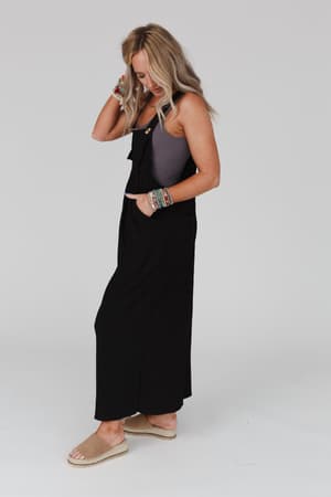 A woman with long blonde hair wears The Nest Serenity Wide Leg Jumpsuit in black over a gray tank top, paired with sandals and colorful bracelets. She stands against a plain light background, looking down and touching her hair.