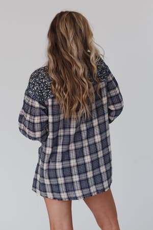 A woman with long, wavy blonde hair faces away from the camera, wearing the Better Together Mixed Print Top in blue gray with plaid and floral patterns on the shoulders and sleeves. The background is plain and light-colored.
