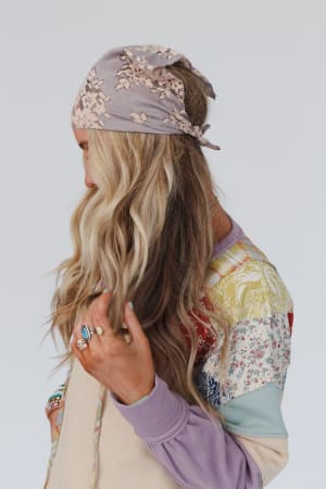 A woman with long, wavy blonde hair wears the Sarah Floral Lace Headscarf in gray and a colorful patchwork-patterned sweater, facing left and looking down as her hand gently touches her hair.