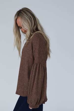 A woman with long blonde hair in dark pants stands in profile against a plain light background, smiling gently while wearing The Nest Sweet Heart Balloon Sleeve Knit Sweater - Mocha.