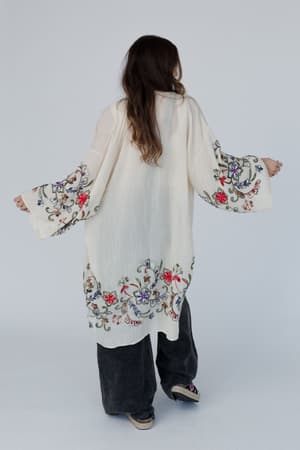 A person with long hair, seen from behind, wears the Fun In Florals Kimono - Cream, featuring sheer fabric and embroidered floral designs on the sleeves and hem, styled with wide-leg dark pants and platform shoes.