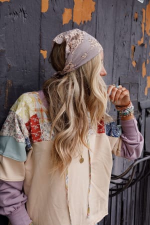 A woman with long wavy blonde hair wears the Sarah Floral Lace Headscarf in gray, a patchwork sweater, and colorful beaded bracelets while standing sideways against a weathered wooden wall with peeling paint.