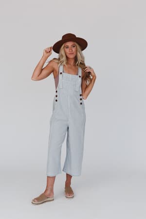 A woman in a brown wide-brim hat, boho-chic beige slip-on sandals, and the High Hopes Denim Overalls - Denim Stripe poses against a white background, holding her hat by the brim with one hand.