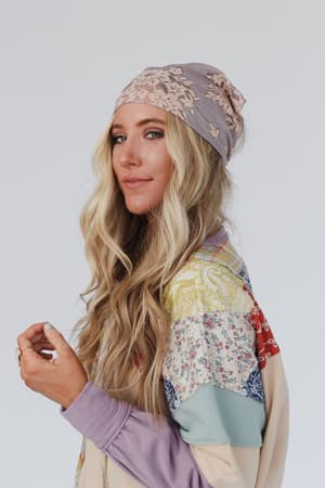 A woman with long blonde wavy hair wears the Sarah Floral Lace Headscarf in gray and a colorful patchwork jacket, smiling softly at the camera against a plain light background.