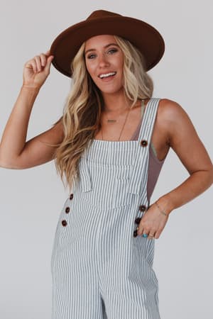 A smiling woman with long blonde hair wears a brown hat, a lavender top, and the High Hopes Denim Overalls - Denim Stripe. She holds her hat's brim with one hand and has the other in her pocket, posing against a light background.