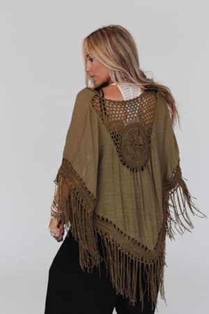 A woman with long blonde hair models the Dreamy Macramé Kimono in olive, featuring bohemian crochet and tassel fringe, layered over a white top and black pants against a light background.