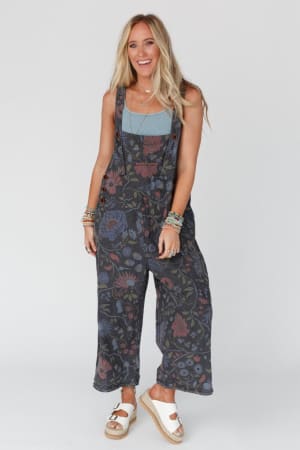 A smiling woman with long blonde hair wears the High Hopes Slouchy Denim Overalls in Floral Black Denim over a blue tank top, accessorized with layered jewelry and white platform sandals, against a plain white background.