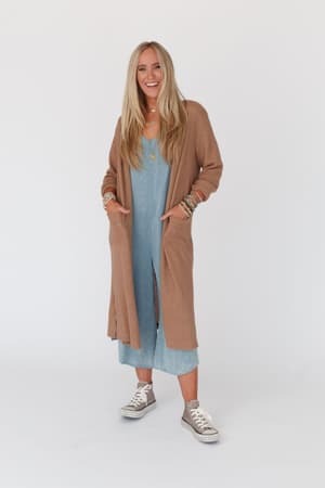 A woman with long blonde hair smiles against a white background, wearing a light blue dress, layered necklaces, bracelets, gray sneakers, and The Nest Jayde Waffle Knit Cardigan in taupe with her hands in its pockets.