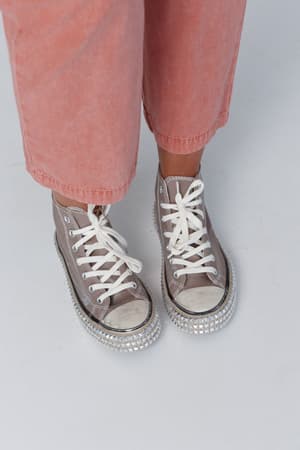 A person wearing light pink pants and the Chantel Studded High Top Sneaker in mocha with white laces and rhinestone detail stands on a white surface.