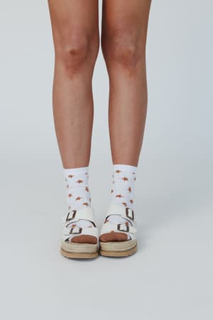 A person wearing white sandals with cork soles and Super Star Socks - White, featuring orange stars, stands against a plain light background. Only the lower legs and feet are visible.