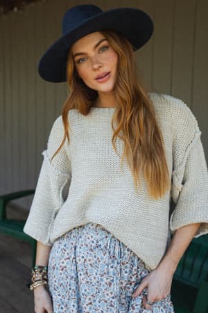 A woman with long red hair wears the Crisp Air Sweater in oatmeal, paired with a wide-brimmed navy hat and a floral skirt. She stands outdoors, looking at the camera with a relaxed expression.