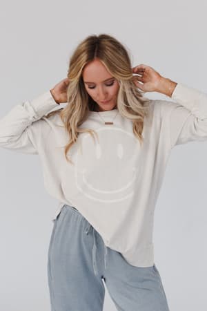 A woman with blonde wavy hair wears the Catch My Smile Pullover Top in khaki and light blue drawstring pants, posing with her hands near her head against a plain light background.