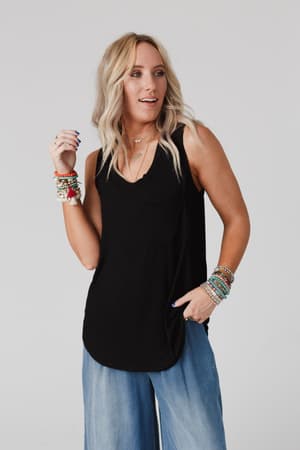 A woman with wavy blonde hair wears The Nest Shelli Scoop Neck Tank Top in black, paired with light blue wide-leg jeans. She accessorizes with layered necklaces and colorful beaded bracelets, standing against a plain light background.