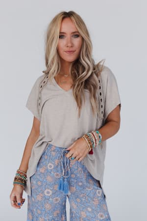 A woman with long blond hair wears the Camden Lace V Neck Top in camel and blue floral pants. She accessorizes with colorful beaded bracelets and layered necklaces, standing against a plain light background.
