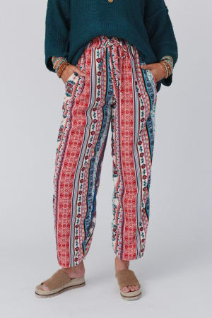 A person stands with hands in their pockets, wearing The Nest Staycation Wide Leg Drawstring Pant in Red Clay, a dark green sweater, brown bracelets, and tan sandals against a plain light background.