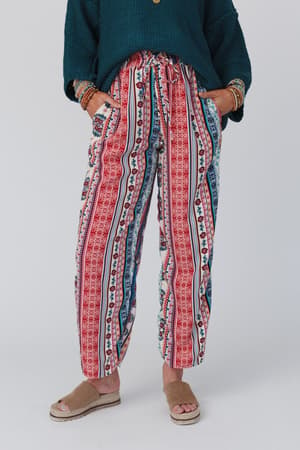 A person stands with hands in their pockets, wearing The Nest Staycation Wide Leg Drawstring Pant in Red Clay, a dark green sweater, brown bracelets, and tan sandals against a plain light background.