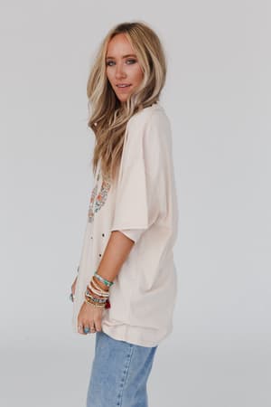 A woman with long blonde hair wears the Jude Oversized Peace Sign Graphic Tee in Oatmeal with light blue jeans and colorful bracelets, standing sideways against a white background and smiling softly.