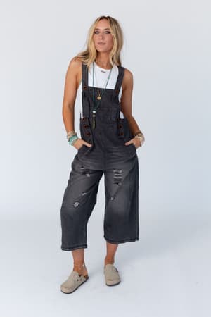 A woman stands against a plain background wearing High Hopes Slouchy Pocket Denim Overalls – Black Pockets over a white tank top, accessorized with bracelets, necklaces, and tan slip-on shoes, hands in her pockets and a relaxed expression.