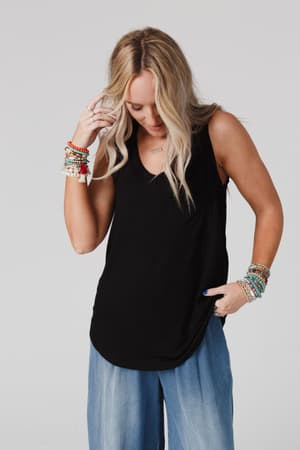 A woman with long blonde hair wears The Nest Shelli Scoop Neck Tank Top in black and wide-leg light blue jeans. She looks down, touching her hair, accessorized with colorful beaded bracelets against a plain light gray background.
