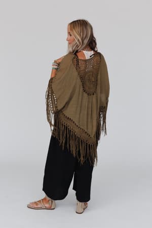A woman faces away, wearing the Dreamy Macramé Kimono in olive with a crochet border and tassel fringe, paired with black wide-leg pants, sandals, and beaded bracelets. The kimono’s boho crochet back pops against the plain white backdrop.