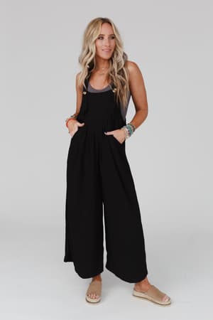 A woman with long blonde hair wears The Nest Serenity Wide Leg Jumpsuit in black and beige sandals, posing against a plain light background. She accessorizes with colorful bracelets on both wrists and has her hands in her pockets.