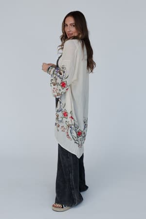 A woman with long brown hair wears the Fun In Florals Kimono - Cream, a sheer, floral-embroidered layer over dark flared pants and platform sandals, posing sideways and looking over her shoulder against a plain light background.