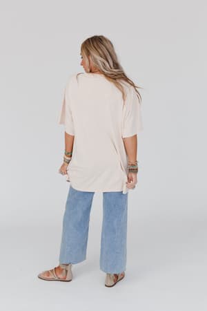 A woman with long blonde hair stands facing away, wearing the Jude Oversized Peace Sign Graphic Tee in oatmeal, paired with blue wide-leg jeans, sandals, and multiple bracelets. The background is plain white.