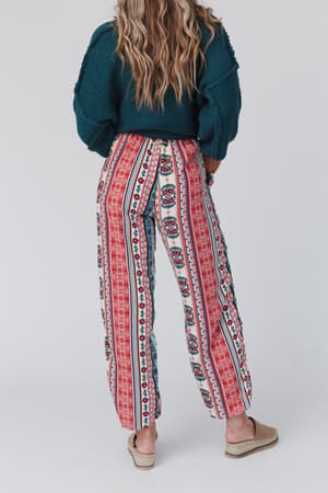 A person with long wavy hair, in a dark green sweater and The Nest Staycation Wide Leg Drawstring Pant - Red Clay, stands facing away. They wear beige slip-on shoes against a plain, light background.