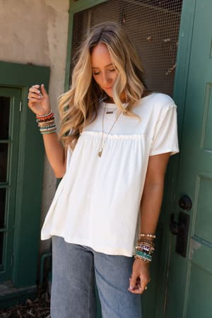 A woman with long blonde hair in loose waves wears the Sunday Morning Babydoll Top - White, styled with blue jeans, colorful beaded bracelets, and a pendant necklace. She stands by a green door, looking down gently.