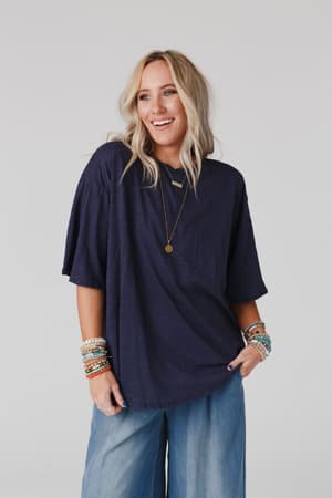 A smiling woman with long blonde hair wears The Nest Your Favorite Slub Tee Shirt in navy, paired with wide-leg jeans, layered necklaces, and colorful beaded bracelets. She poses against a plain light background.
