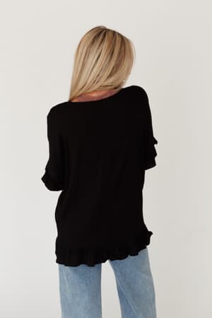 A person with long blonde hair, wearing The Nest Let Me Be Ruffled Top in black and light blue jeans, stands facing away against a plain white background.
