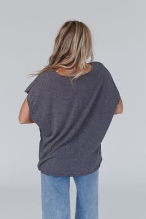 A person with long blonde hair is standing with their back to the camera, wearing The Nest Camden Lace V Neck Top in charcoal and light blue jeans, against a plain light background.
