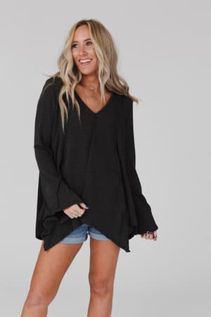 A woman with long blonde hair wears the Golden Hour Long Bell Sleeve Top in charcoal and denim shorts. She smiles, looking to the side, standing against a plain light gray background.