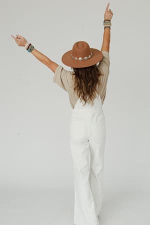 Wearing the Milly Distressed Hem Wide Leg Overall in cream, a person faces away with arms raised and fingers pointing up. Their long, wavy hair flows down and multiple bracelets decorate their wrists.